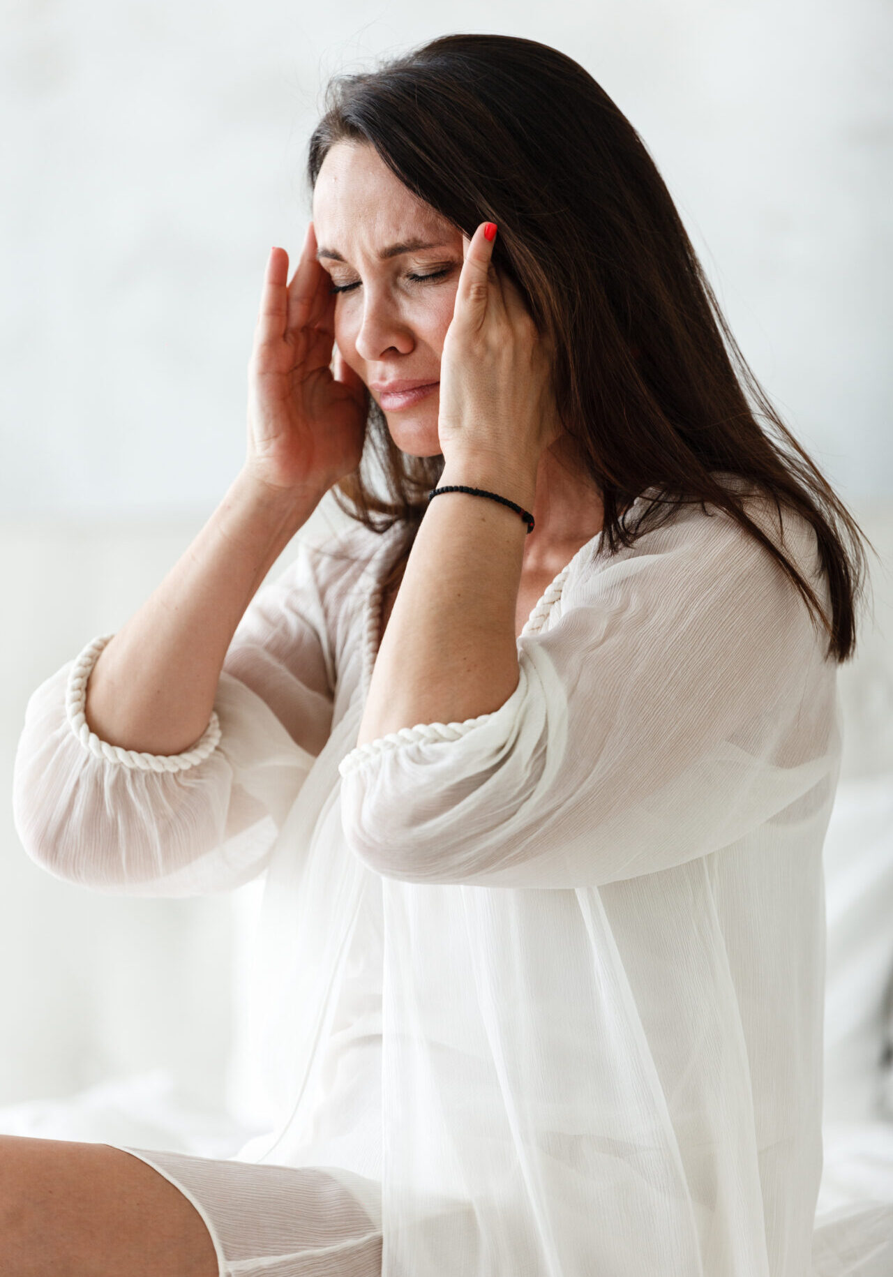 Middle aged woman sitting on a white bed in a bedroom at home touching her head with her hands while having a headache pain and feeling unwell. Suffering from insomnia or migraine.
